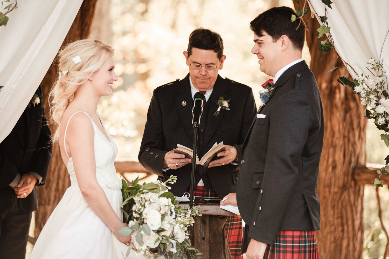 Outdoor wedding ceremony with bride in white gown, groom in tartan kilt, and officiant reading vows under draped arch.