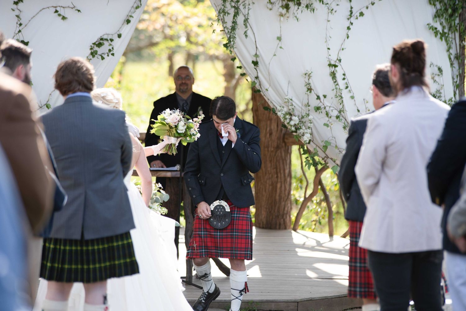 Emotional groom in red tartan kilt bowing head during outdoor wedding ceremony with guests in kilts and floral arch.