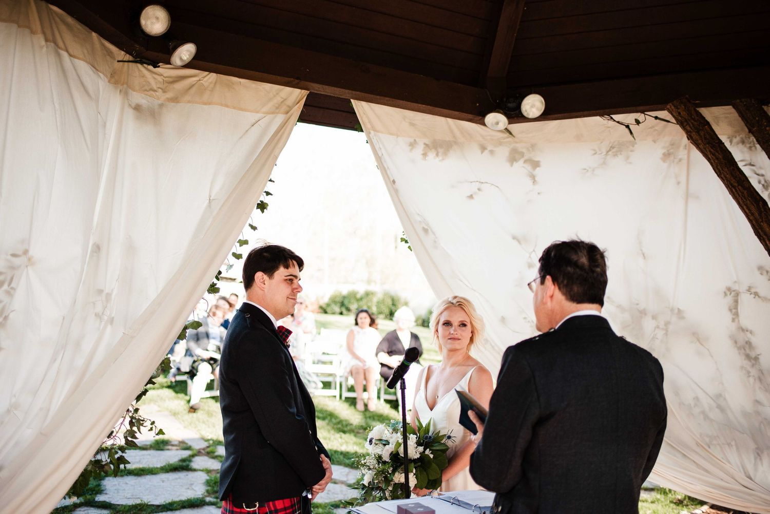 Bride and groom exchange vows under a draped gazebo with string lights during an outdoor wedding ceremony.