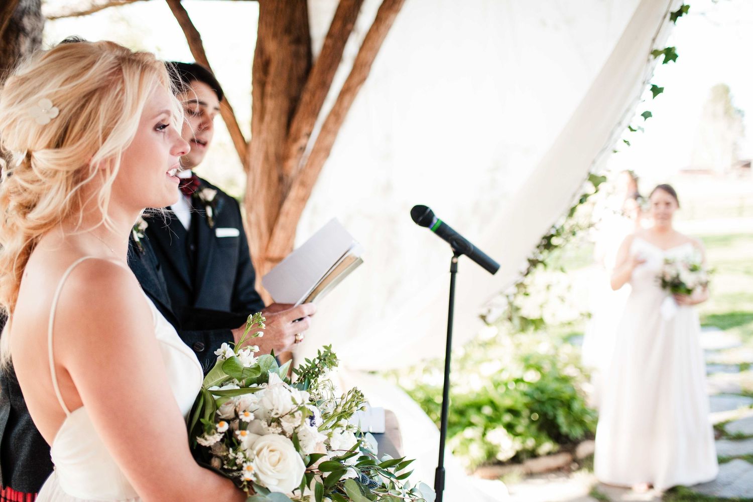 Bride reads vows at outdoor wedding ceremony with officiant and bridesmaid holding white floral bouquet.