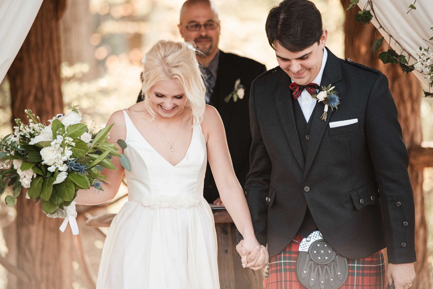 Bride and groom holding hands walking down the aisle, groom wearing a kilt and bow tie at outdoor wedding.