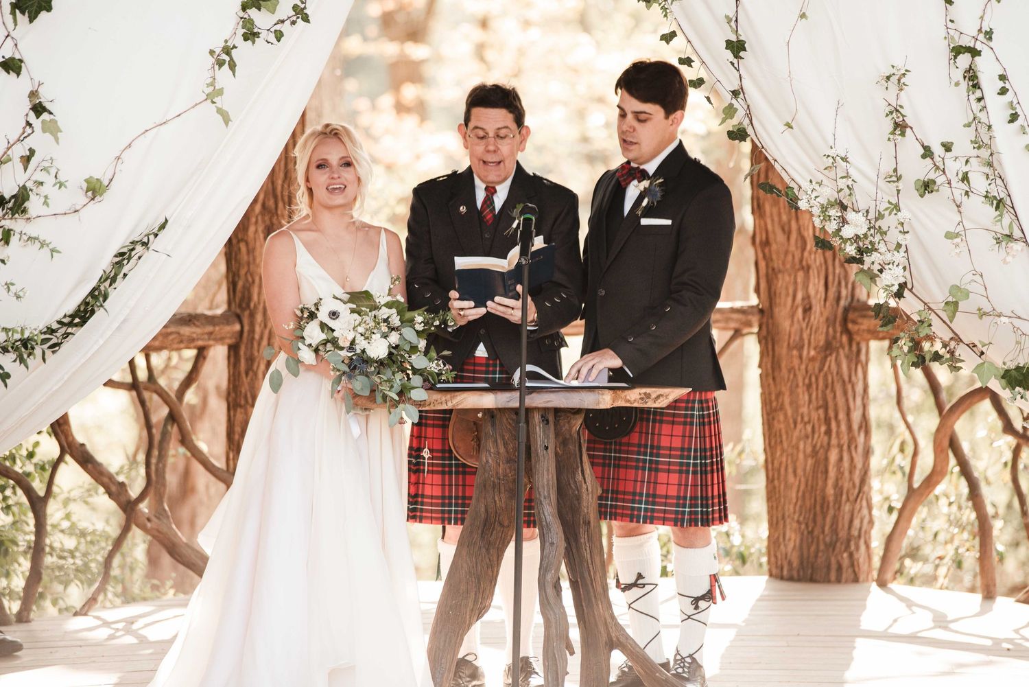 Bride and groom exchange vows at rustic outdoor wedding ceremony with officiant in plaid kilt.
