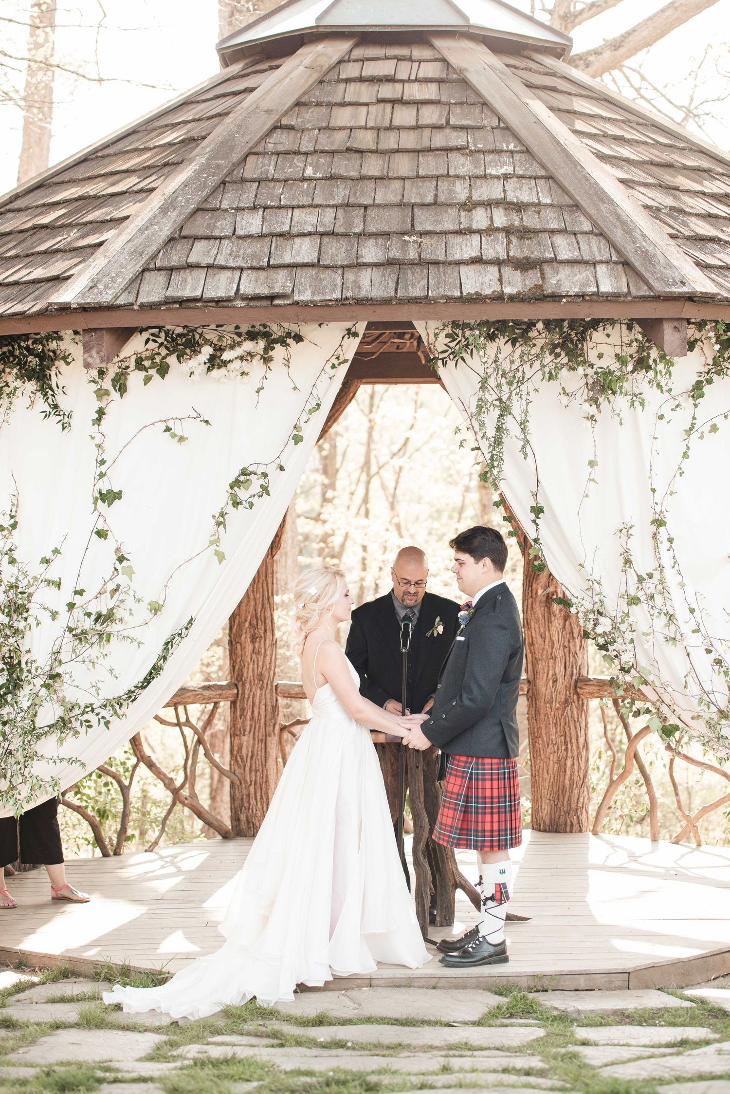 Bride and groom exchange vows under a rustic gazebo draped with white fabric and greenery, groom wearing a tartan kilt.