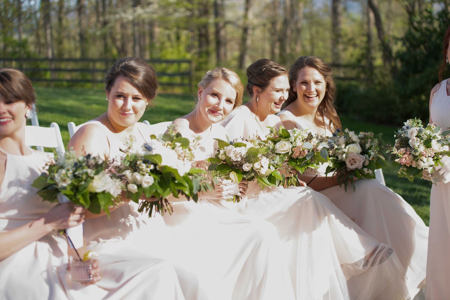 Bridesmaids in white dresses holding lush floral bouquets with greenery at an outdoor wedding ceremony.