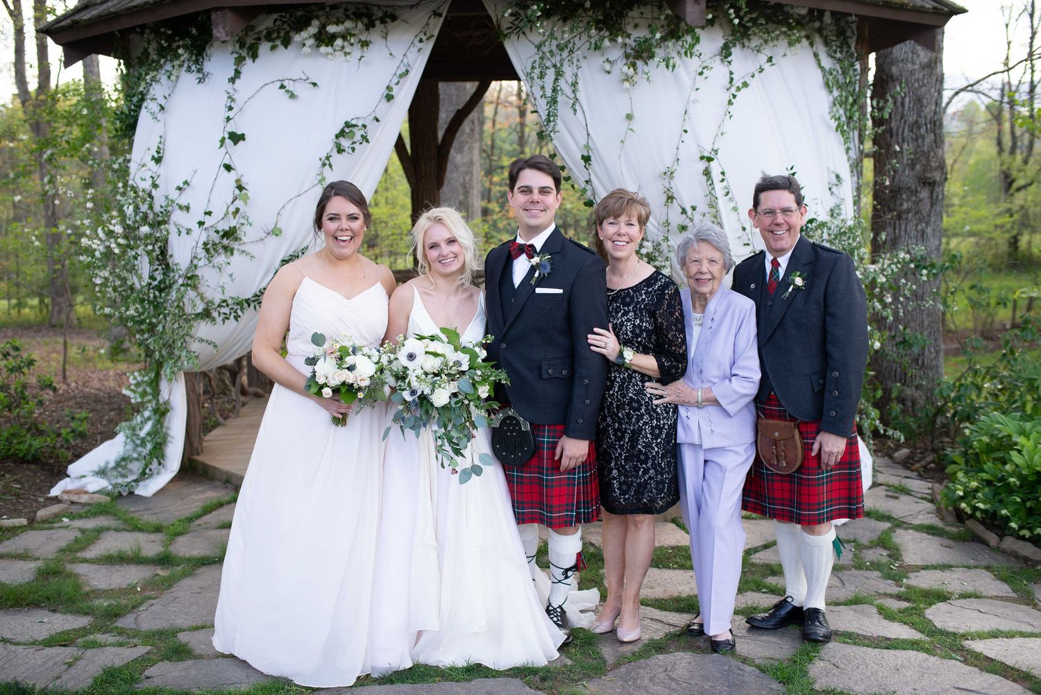 Wedding party poses under floral gazebo, bride in white gown, groom and groomsmen in kilts, outdoors.