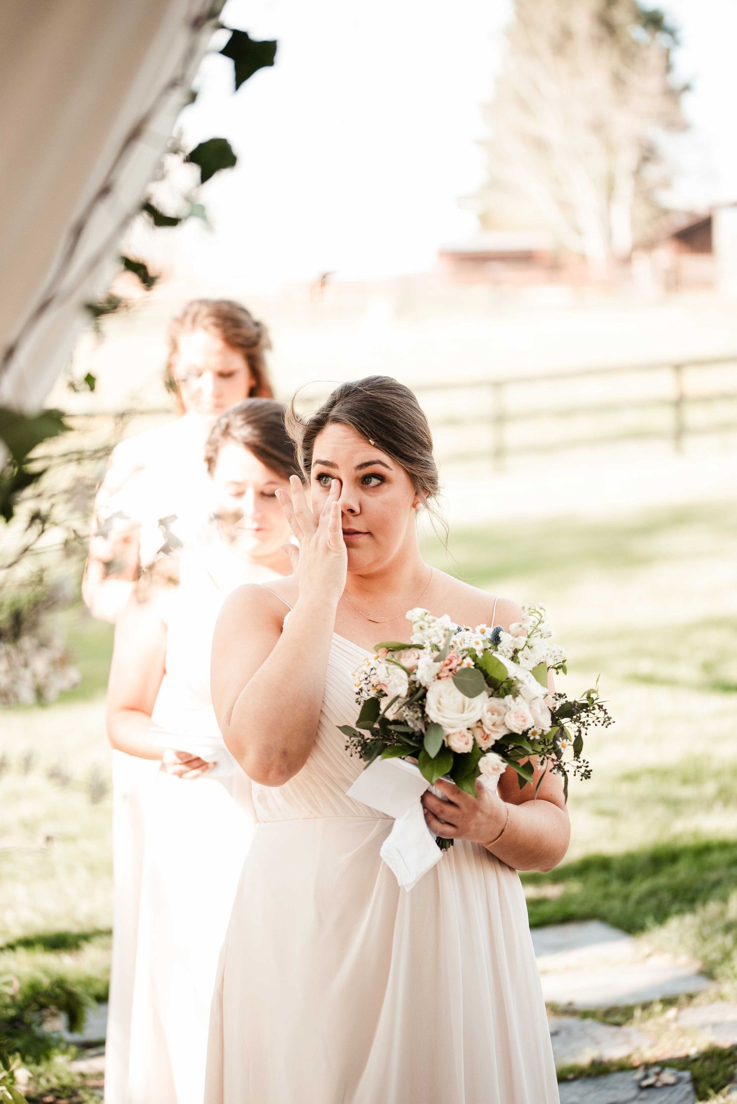 Bridesmaid in white gown wiping tears while holding floral bouquet at outdoor wedding ceremony.