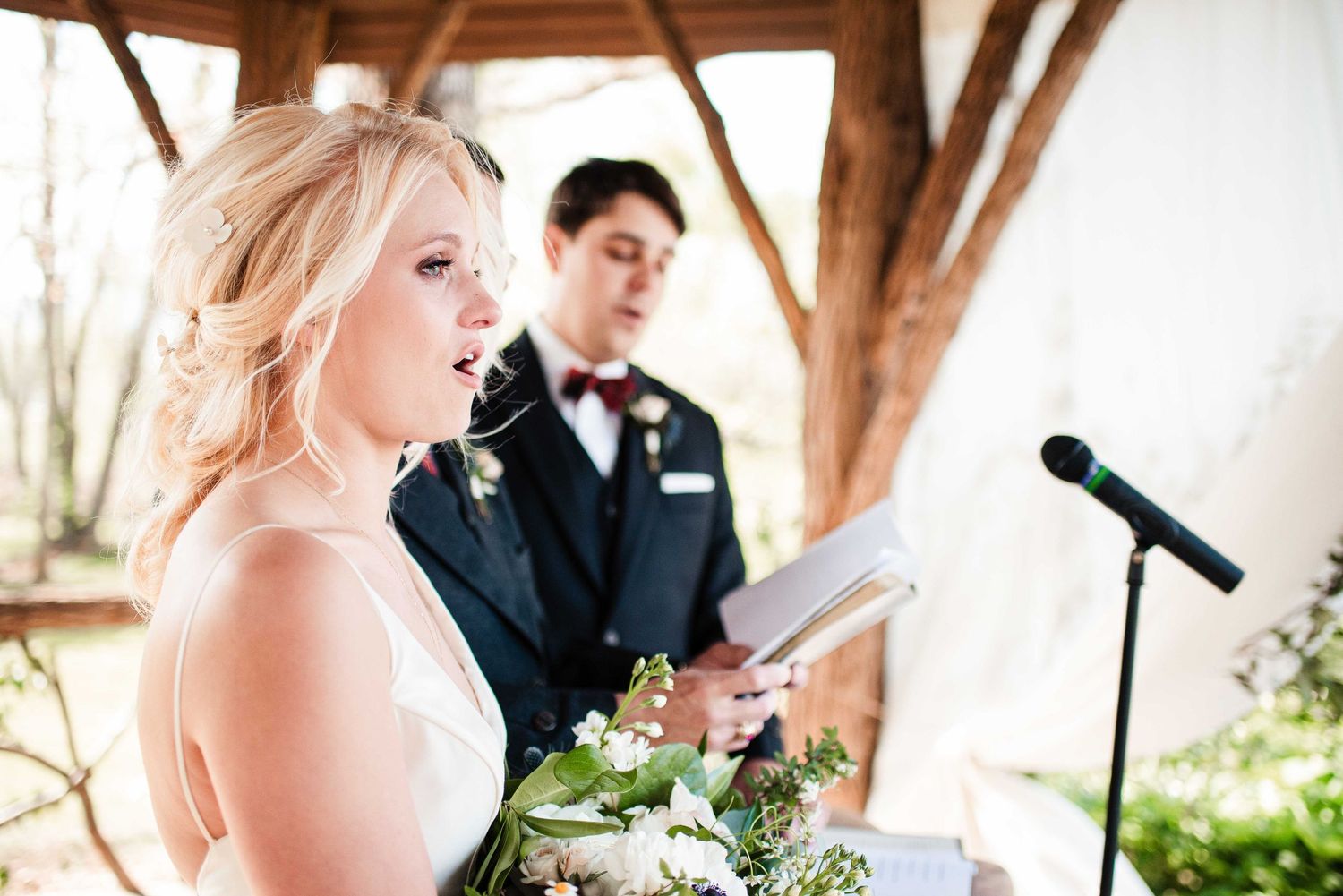 Bride reading vows from book at outdoor wedding ceremony, groom in navy suit with bow tie standing behind her.