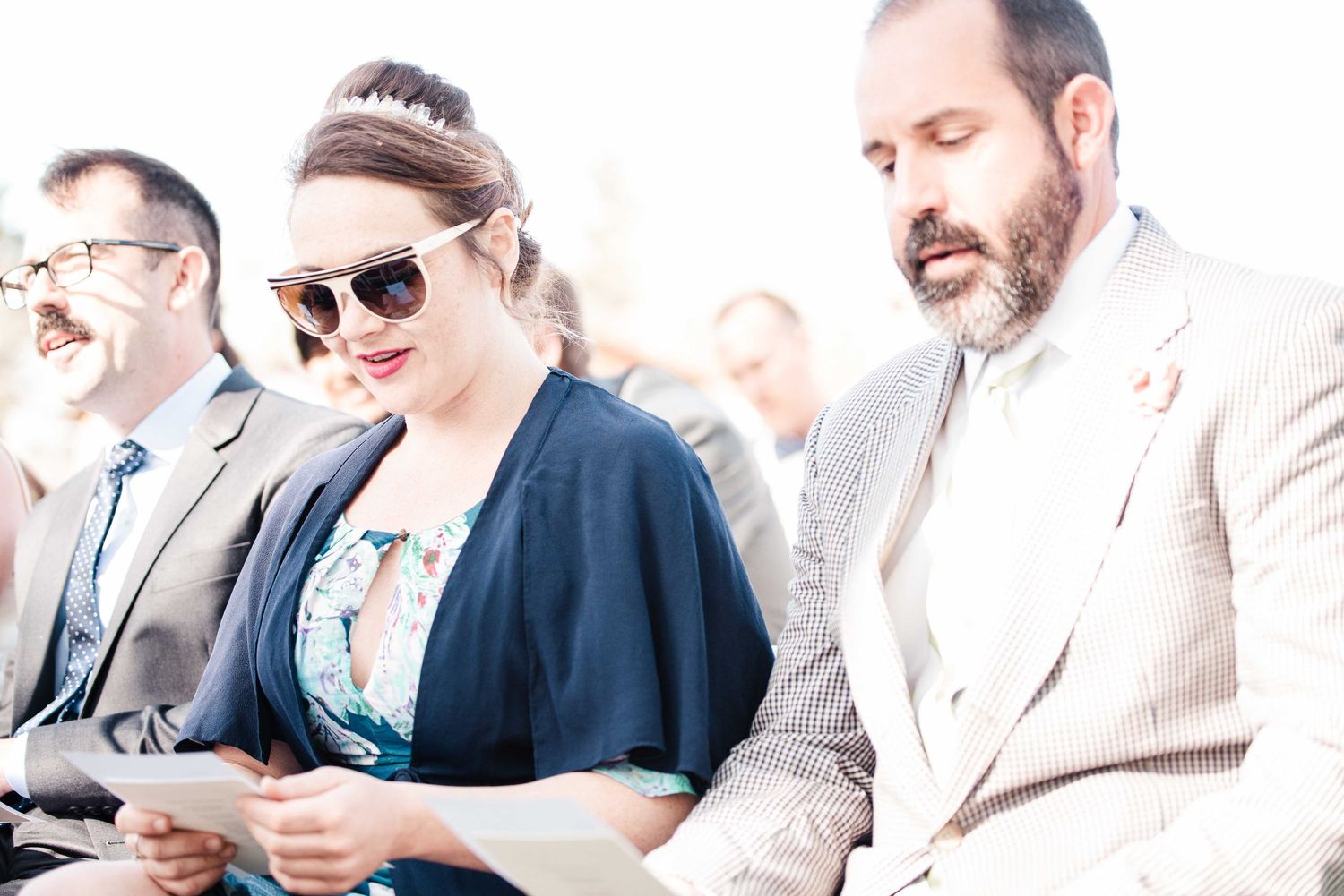 Elegantly dressed wedding guests reading programs at an outdoor ceremony, woman in sunglasses and floral dress.