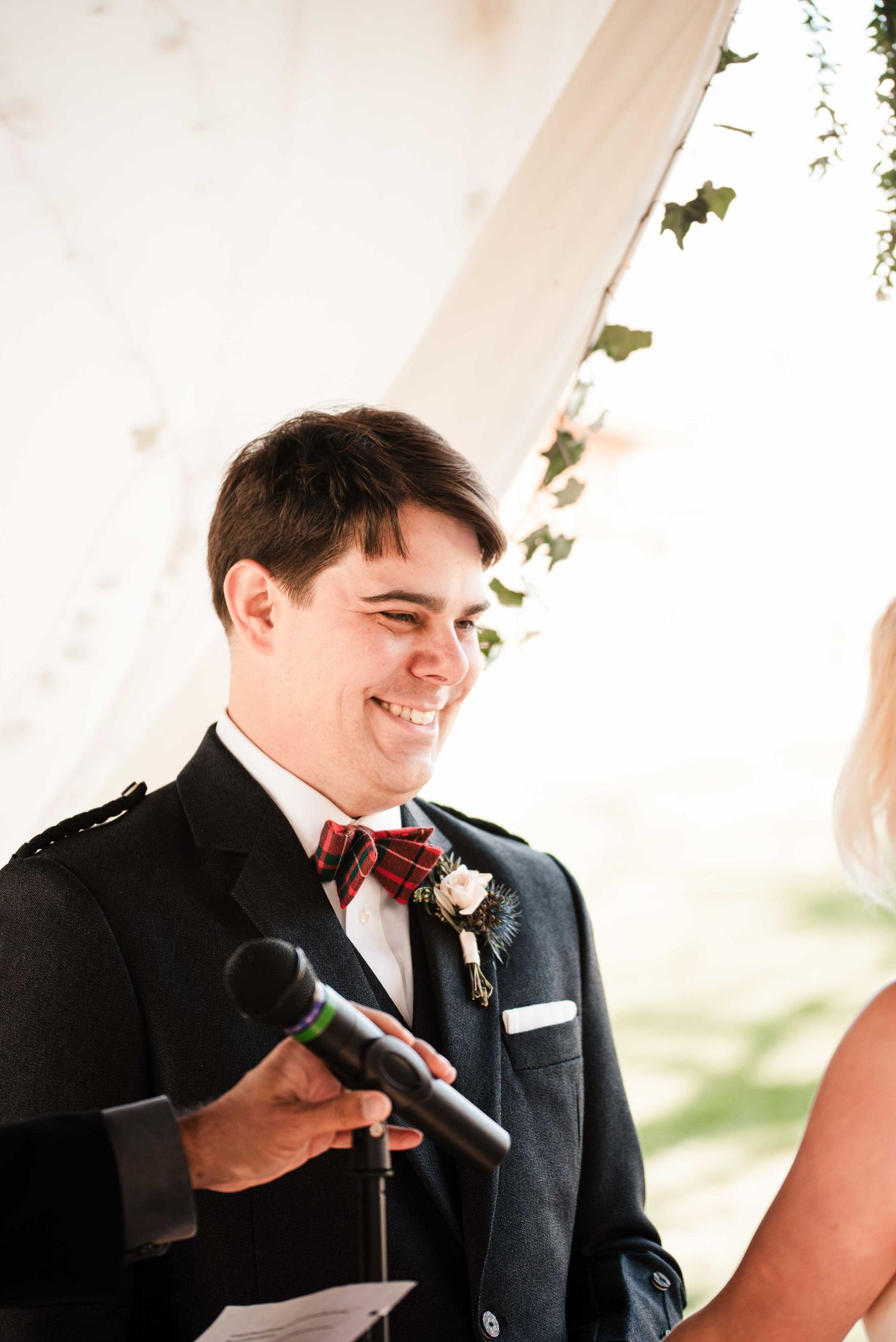 Smiling groom in dark suit with red tartan bow tie and boutonniere holds microphone during outdoor wedding ceremony.