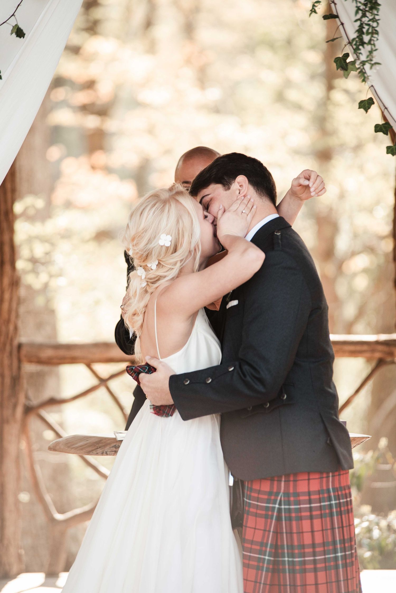 Bride and groom share first kiss at outdoor wedding ceremony, groom wearing tartan kilt and navy blazer.