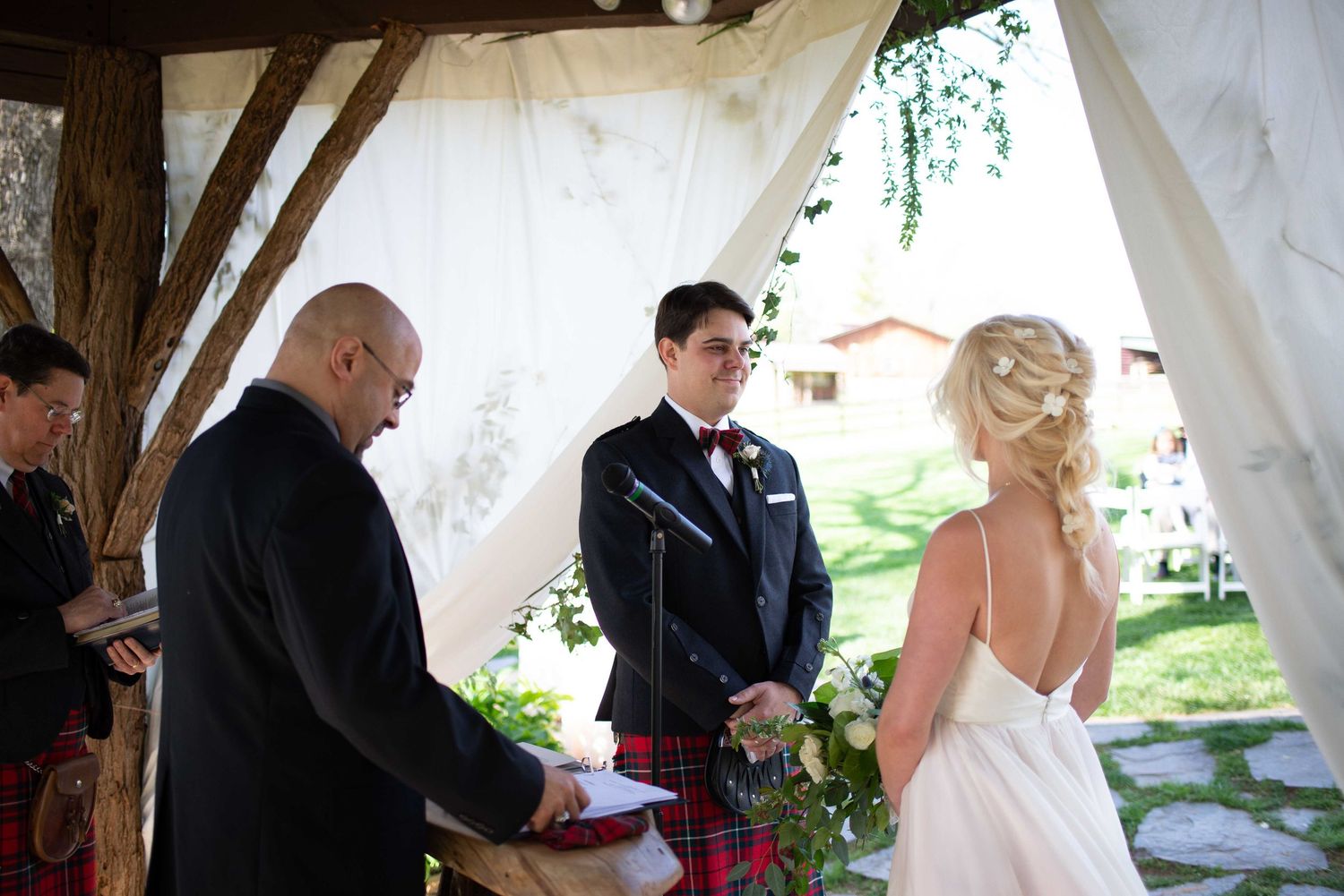 Outdoor wedding ceremony with groom in kilt, bride in white gown, and officiant under draped fabric arbor.