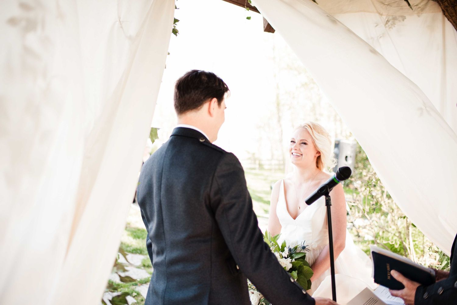 Bride smiling at groom during outdoor wedding ceremony under white tent canopy with microphone stand.
