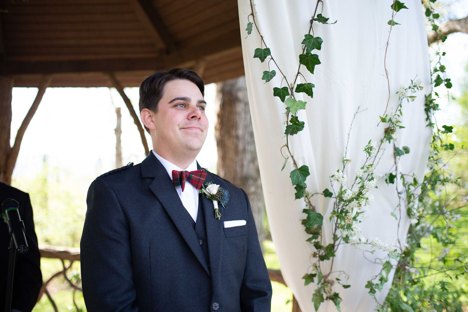 Groom wearing navy suit with burgundy boutonniere smiling emotionally at outdoor wedding ceremony altar.