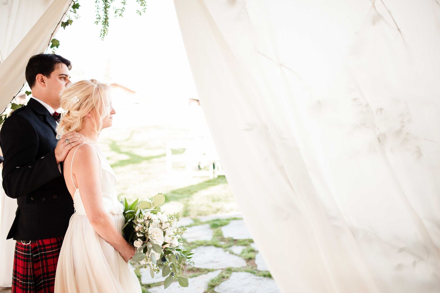 Bride and groom share a romantic moment under flowing white drapes, groom in tartan kilt, bride holding a white bouquet.