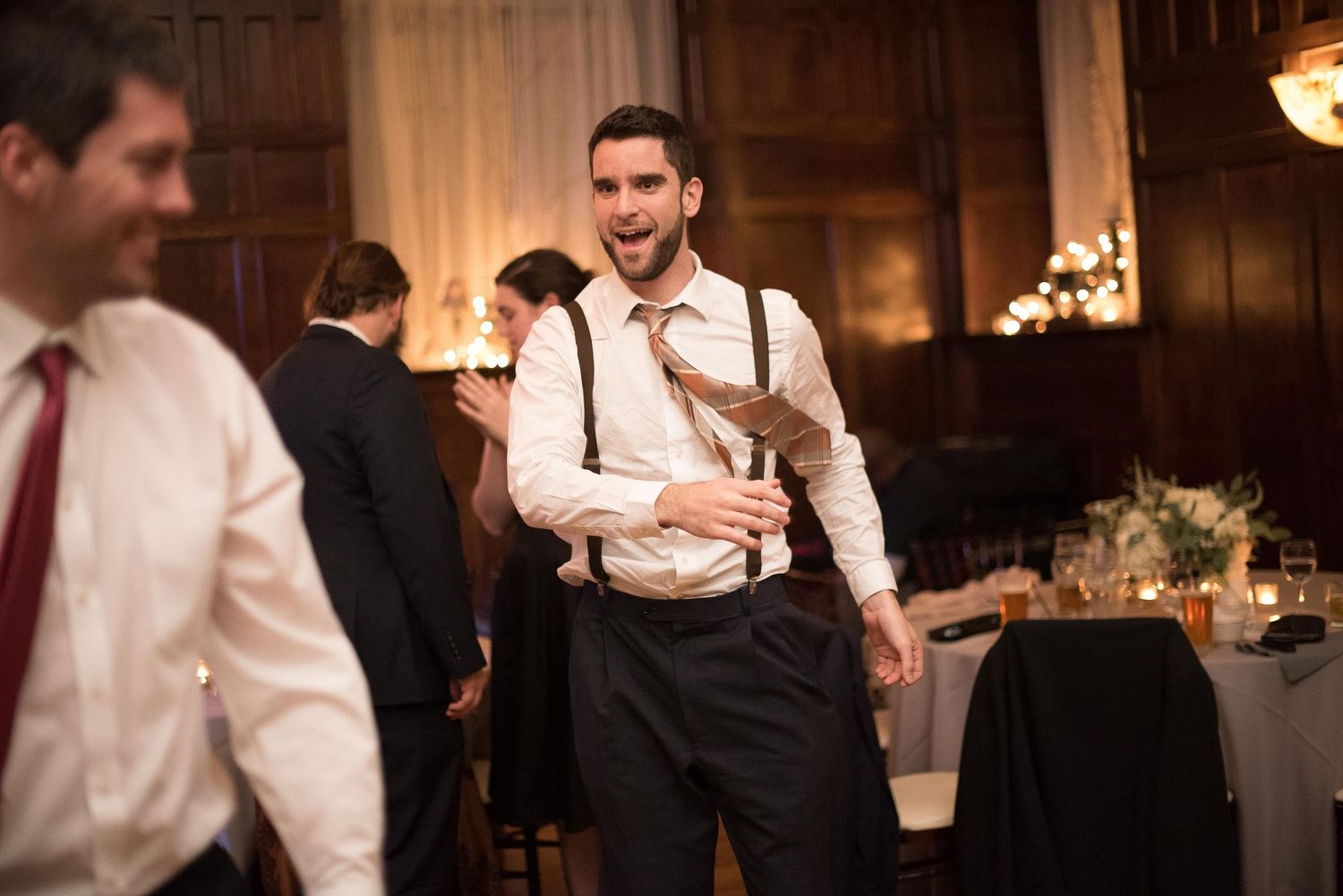 Man in suspenders and tie smiling at a formal wedding reception in an elegant candlelit venue.