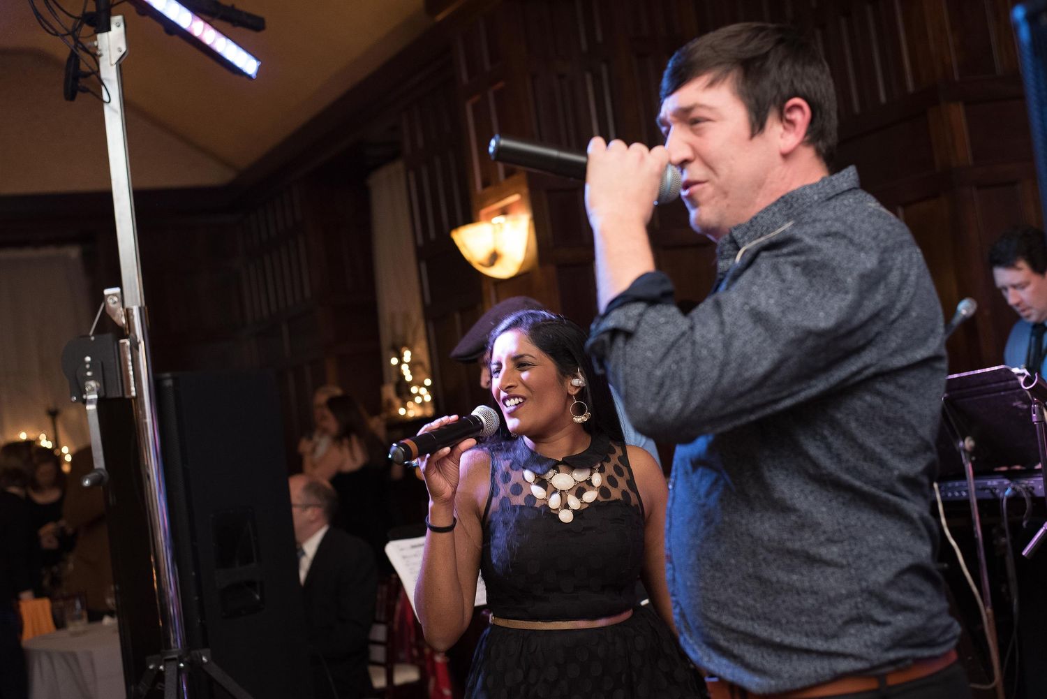 Two singers performing on stage with microphones at an indoor event in a warmly lit venue.