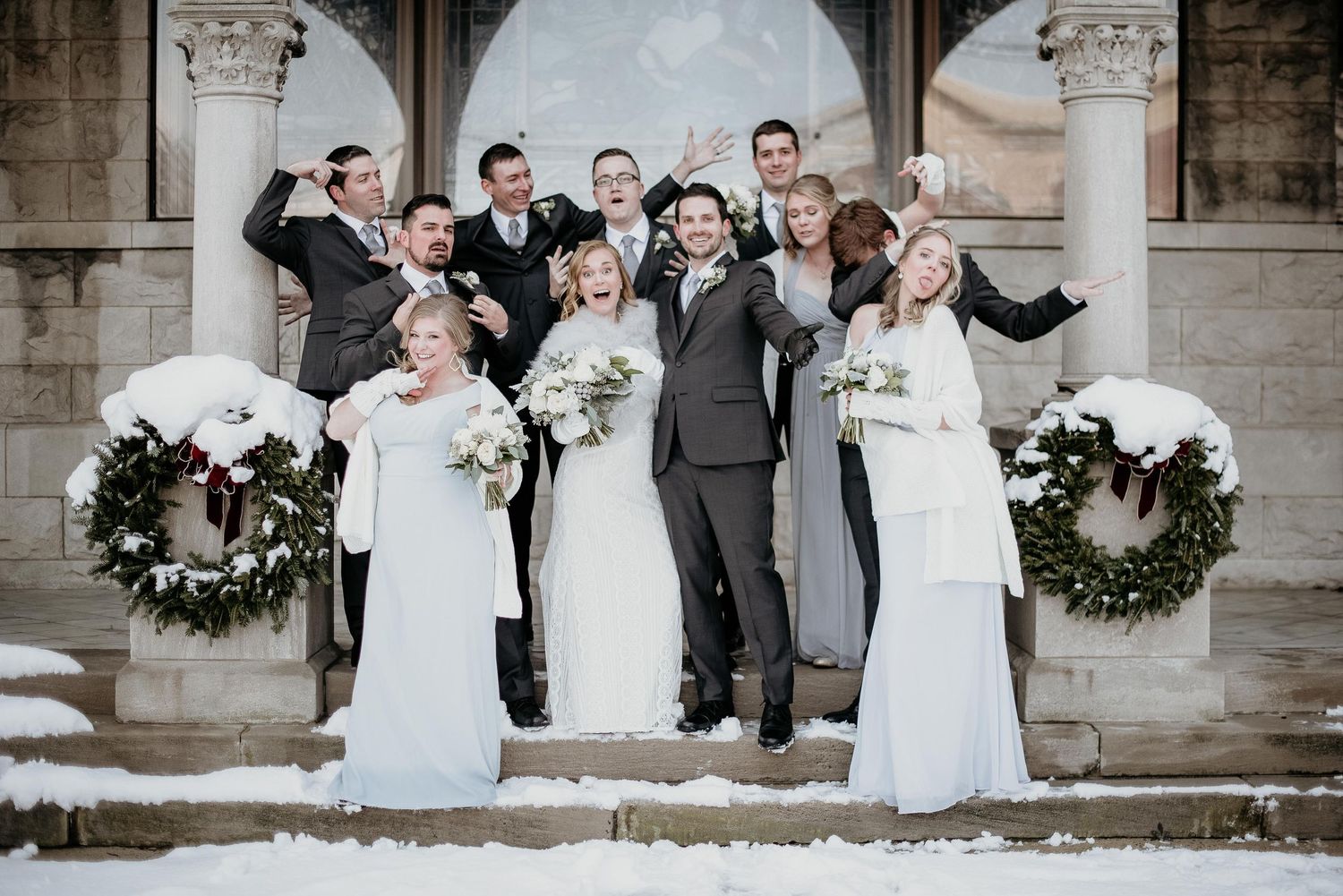 Wedding party celebrating on snowy church steps, bridesmaids in white gowns and groomsmen in gray suits with festive wreaths.