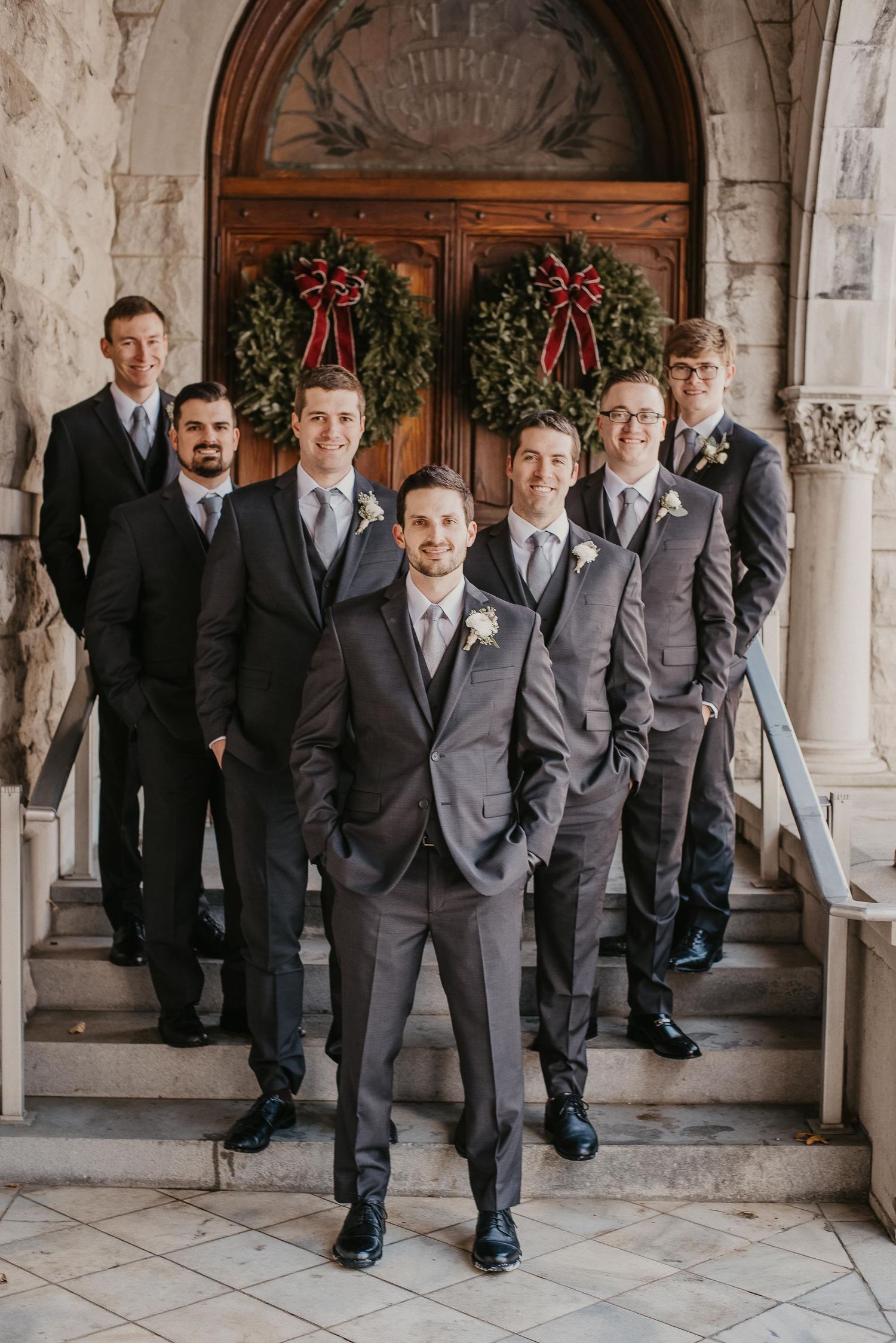 Groom and groomsmen in charcoal suits pose on stone steps before ornate wooden doors adorned with Christmas wreaths.