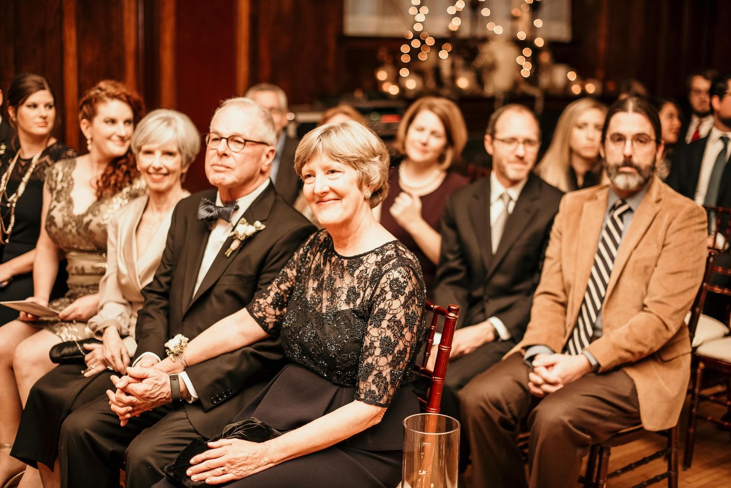 Wedding guests smiling and seated during an indoor ceremony, dressed in formal attire including black lace and suits.