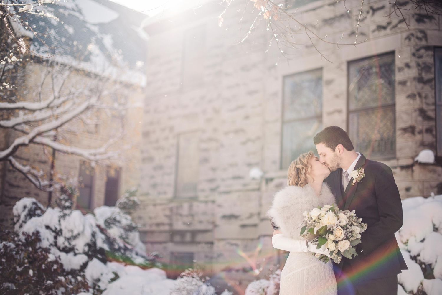 Bride and groom share a romantic kiss in a snowy winter garden, bride holding white bouquet, stone building backdrop.