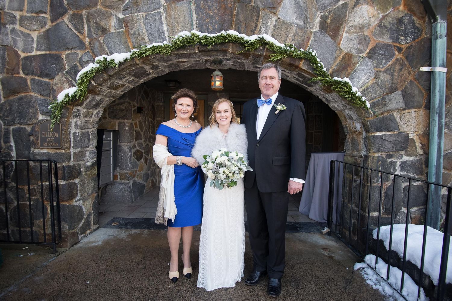 Bride with parents posing in front of stone arch entrance dusted with snow at winter wedding venue.