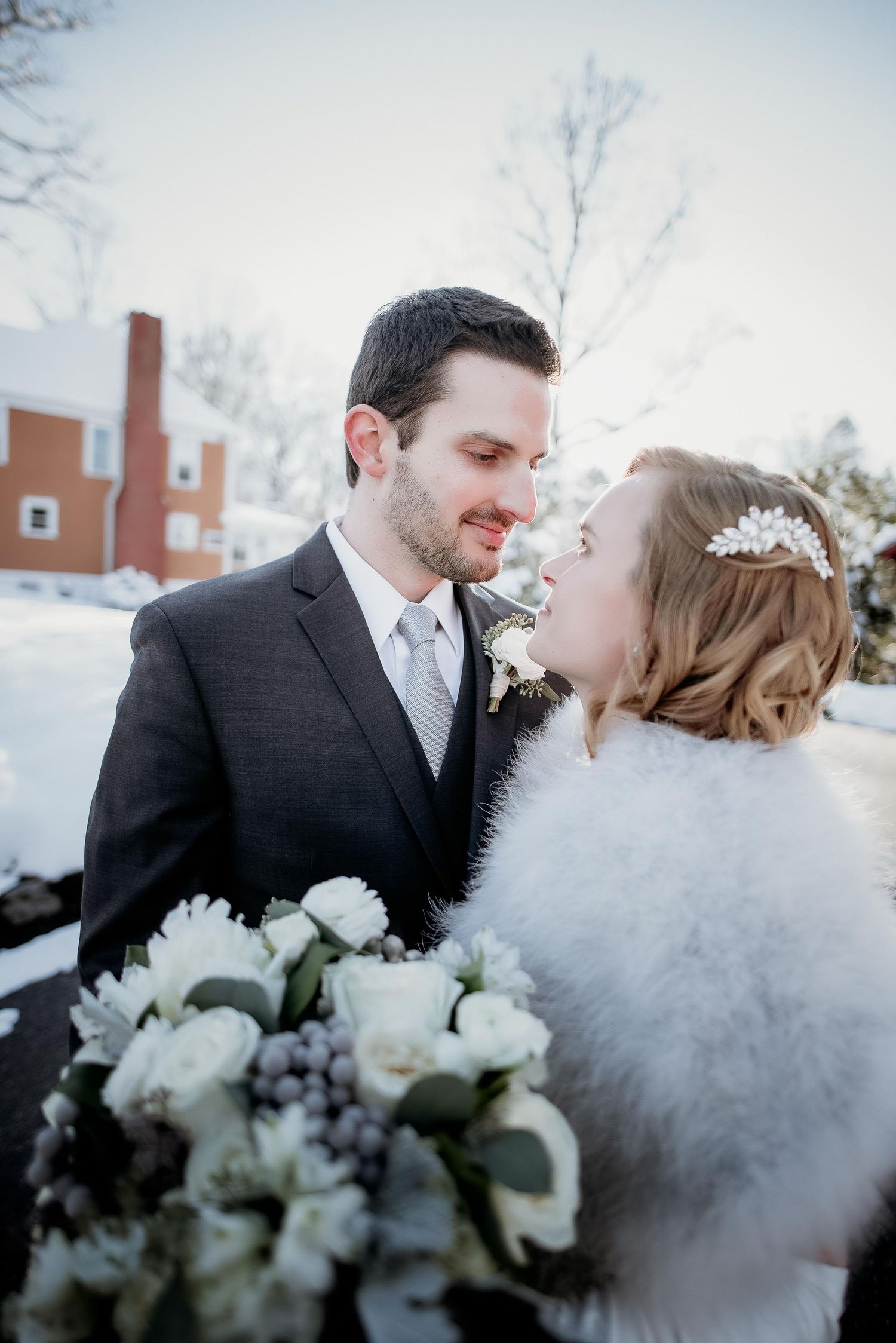 Bride and groom share a romantic winter moment, she holds white bouquet, wearing fur wrap and floral headpiece.
