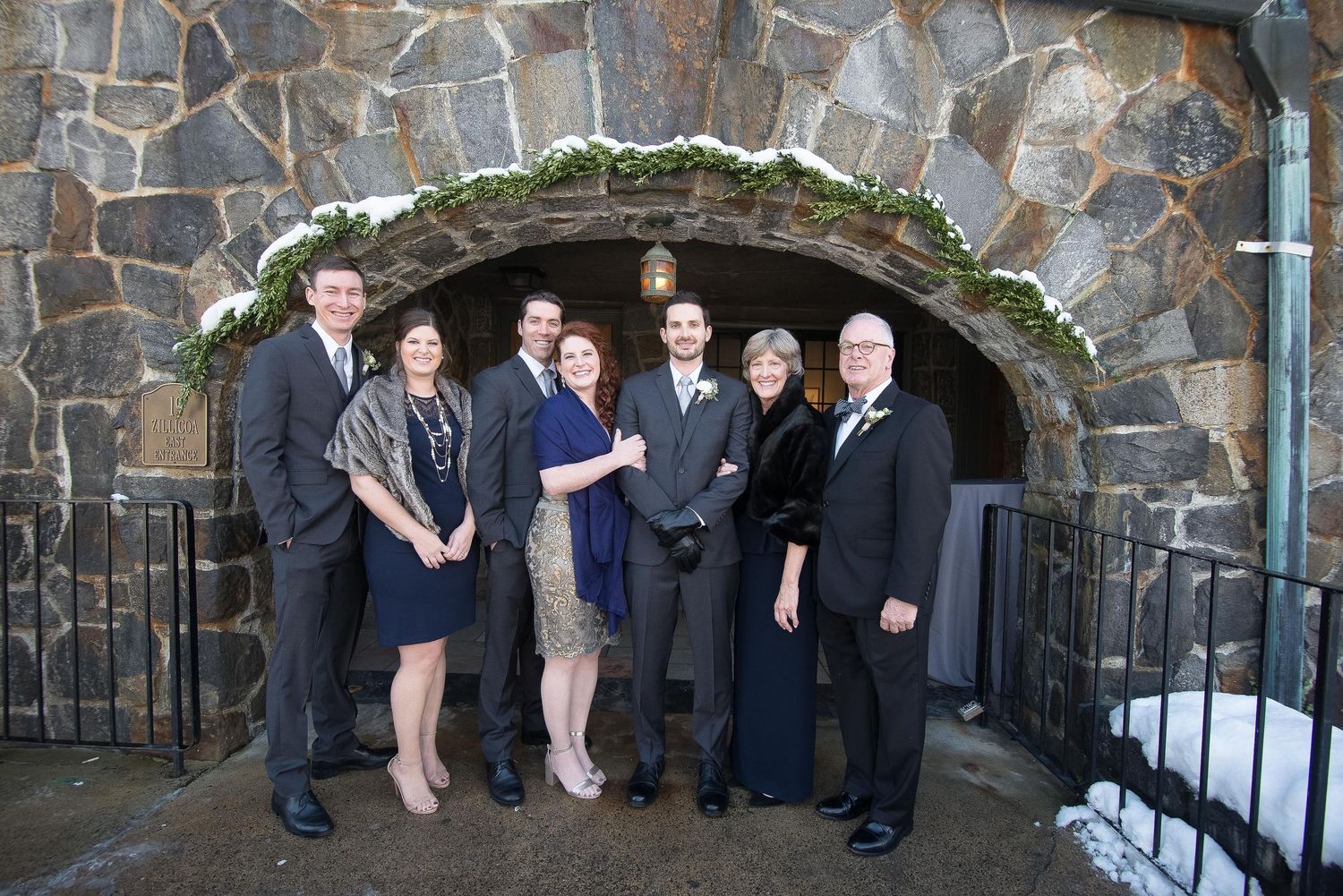 Group of seven people posing together under a stone archway decorated with greenery at a winter wedding.