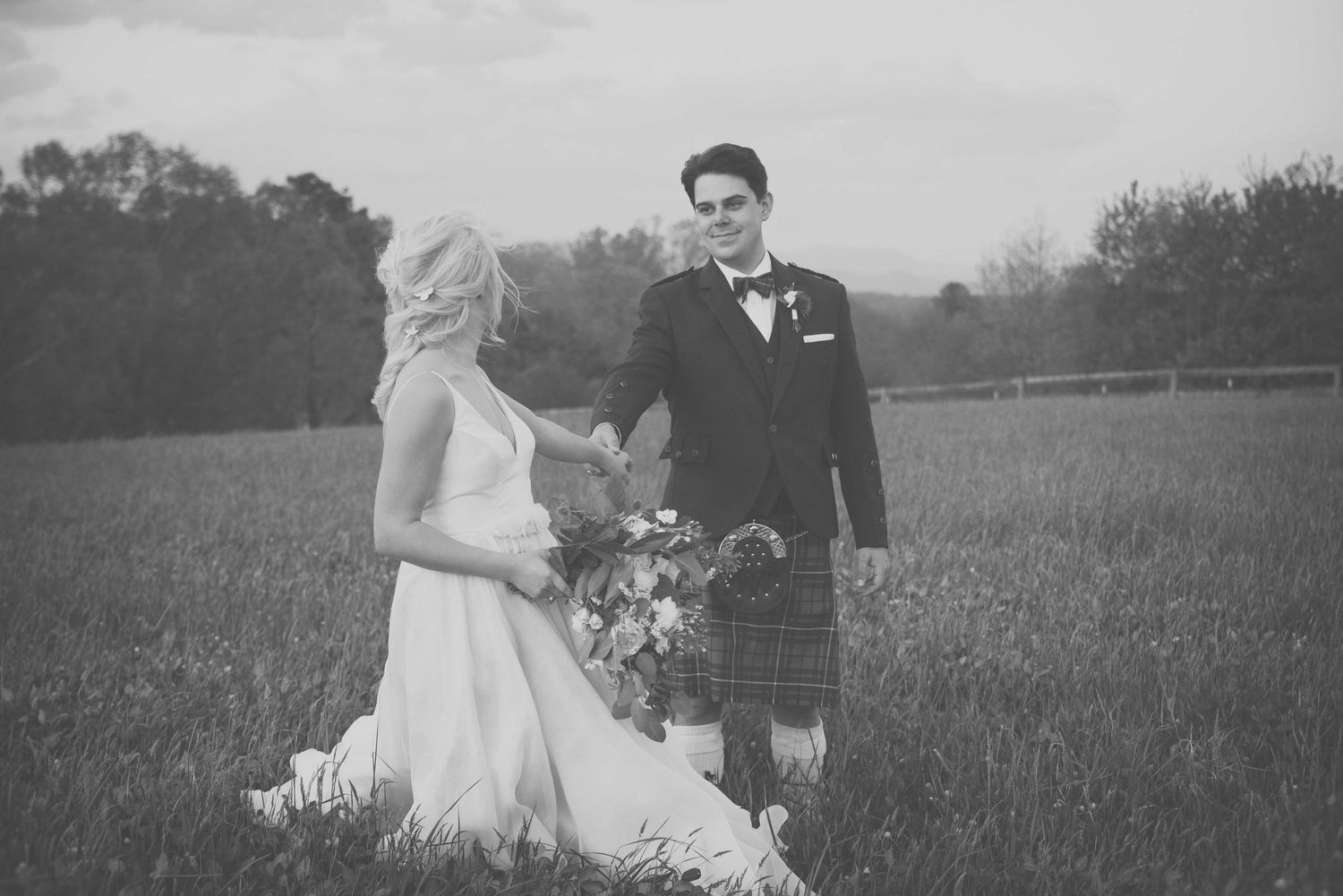 Black and white photo of a bride in white gown and groom in kilt standing in a lush green field.