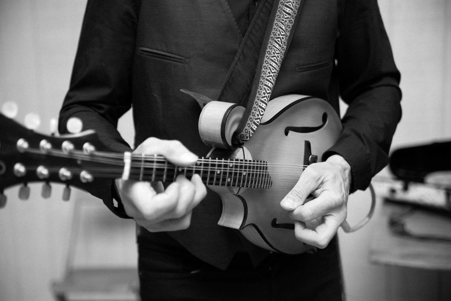 Black and white photo of a musician playing a mandolin, wearing a dark suit with a strap.