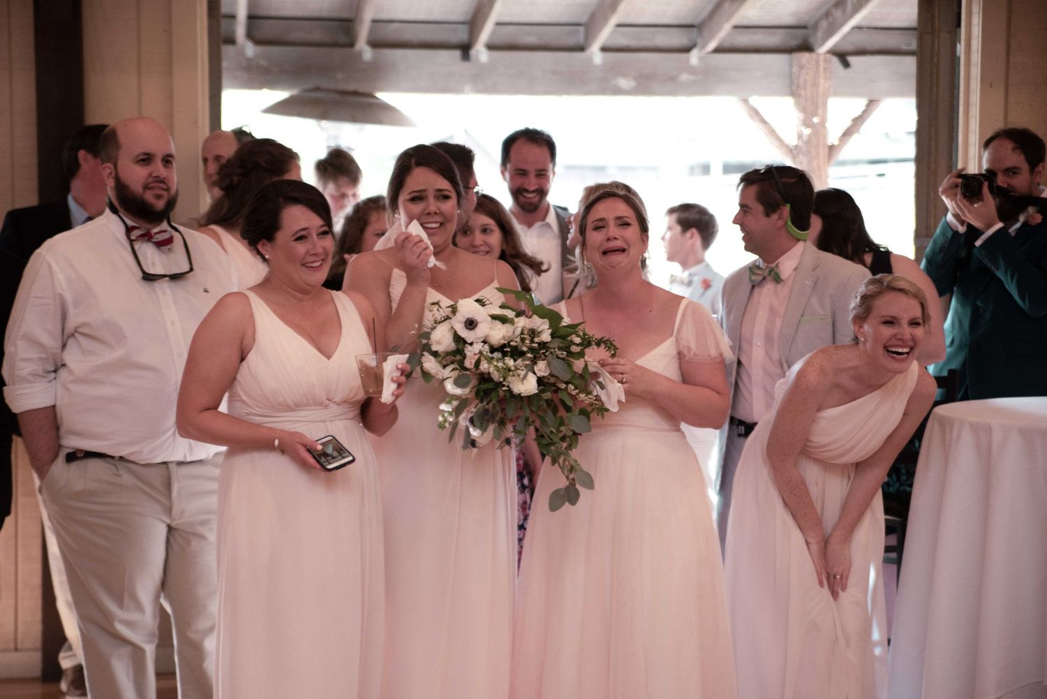 Bridesmaids in blush pink gowns laugh and celebrate at a wedding reception holding a white floral bouquet.