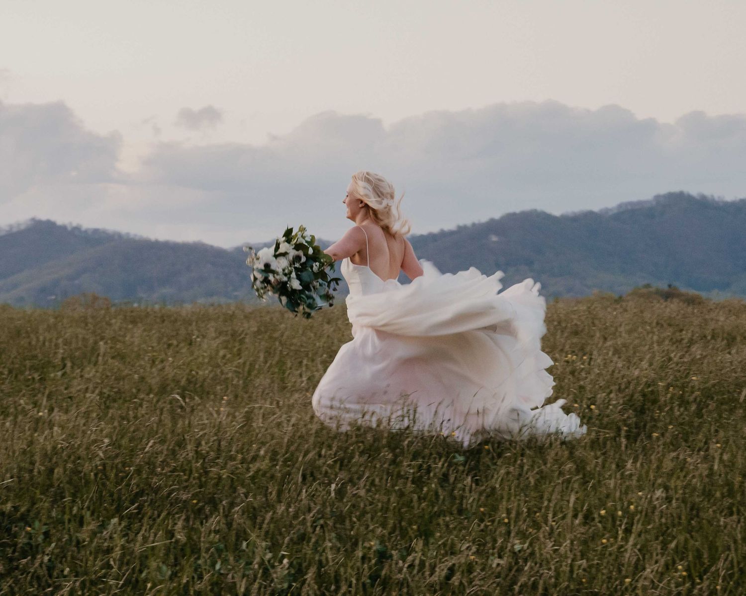 Bride in flowing white gown running through green meadow holding bouquet with mountain backdrop.