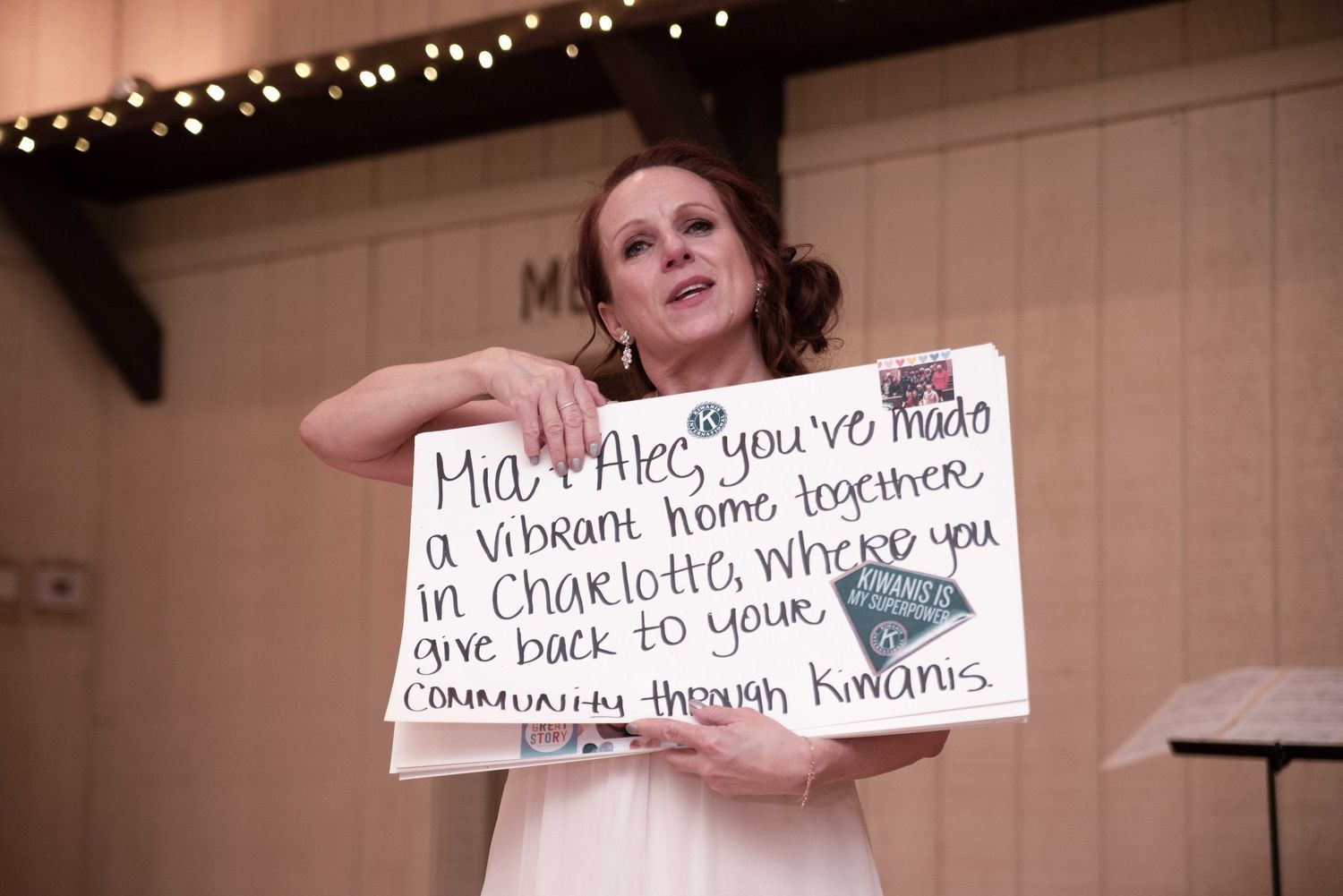 Woman holding a handwritten sign mentioning Mia, Alex, Charlotte, and Kiwanis community at an indoor event.