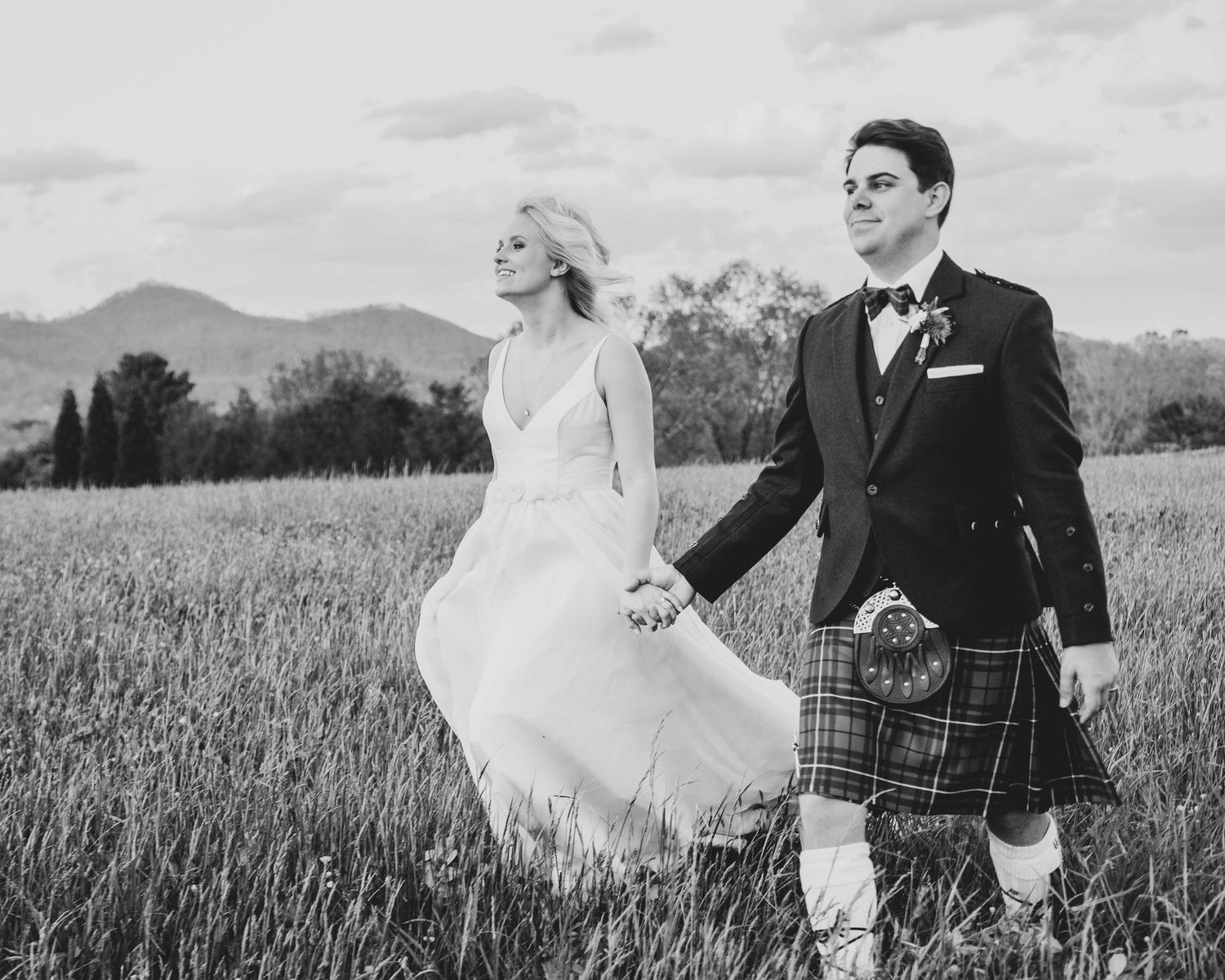 Bride and groom holding hands in a field, groom wearing a Scottish kilt, black and white wedding photo.
