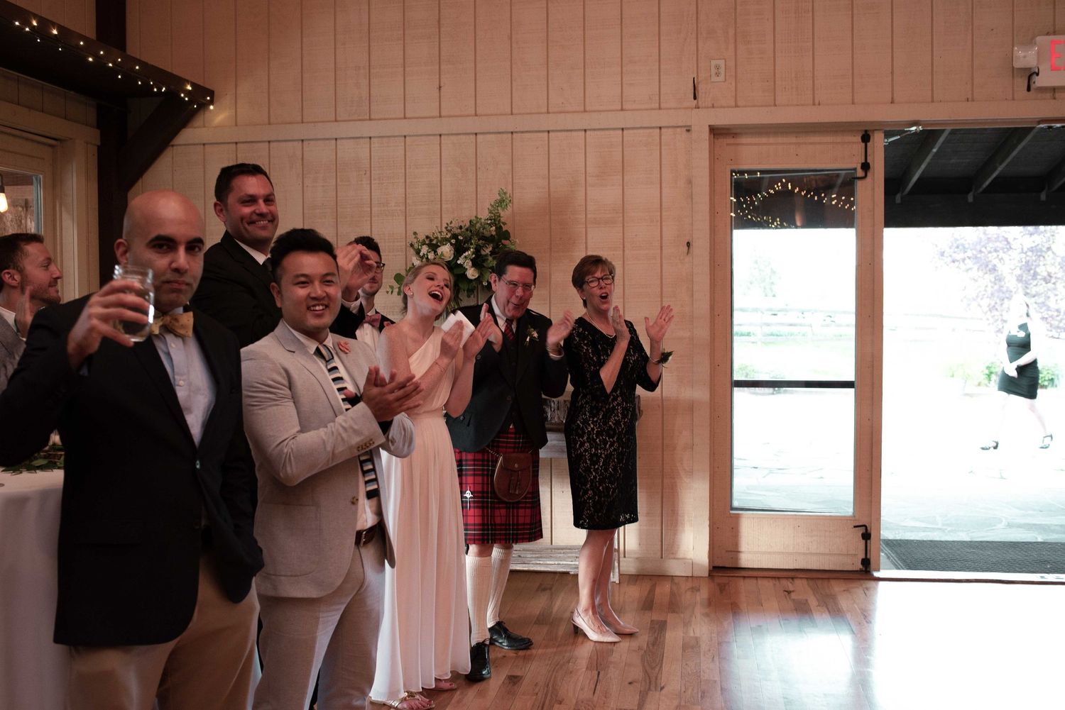 Wedding guests clapping and celebrating indoors at a reception venue with wood-paneled walls and warm lighting.