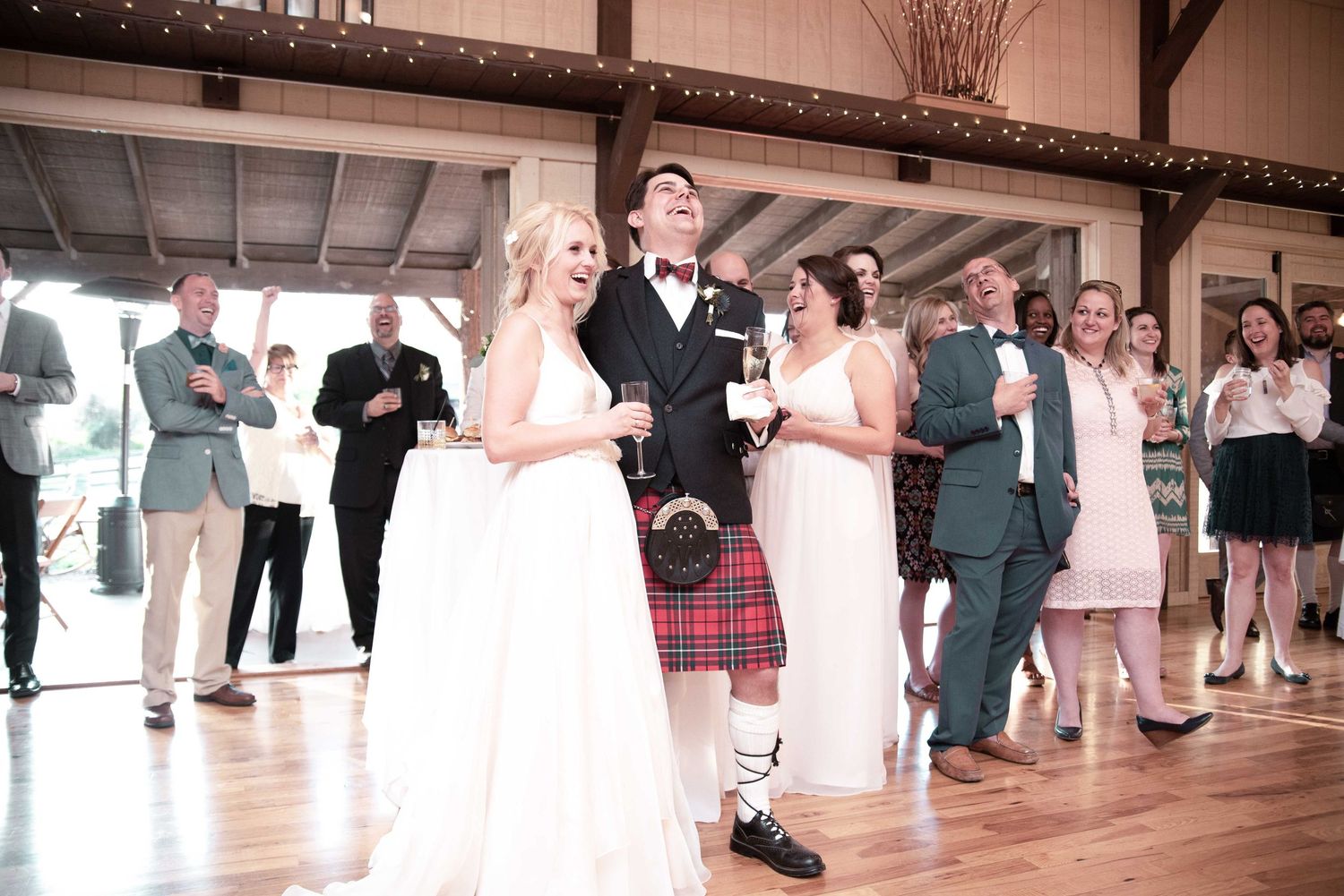 Bride and groom in tartan kilt celebrate at wedding reception in rustic barn venue with guests.
