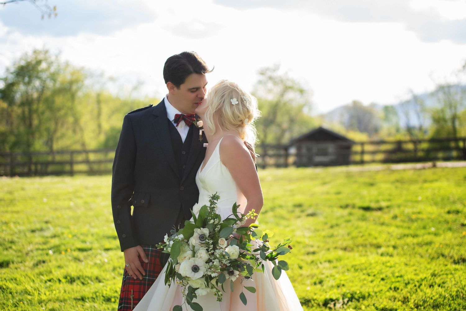 Bride and groom share a romantic moment in a lush green field, bride holding a white floral bouquet with greenery.
