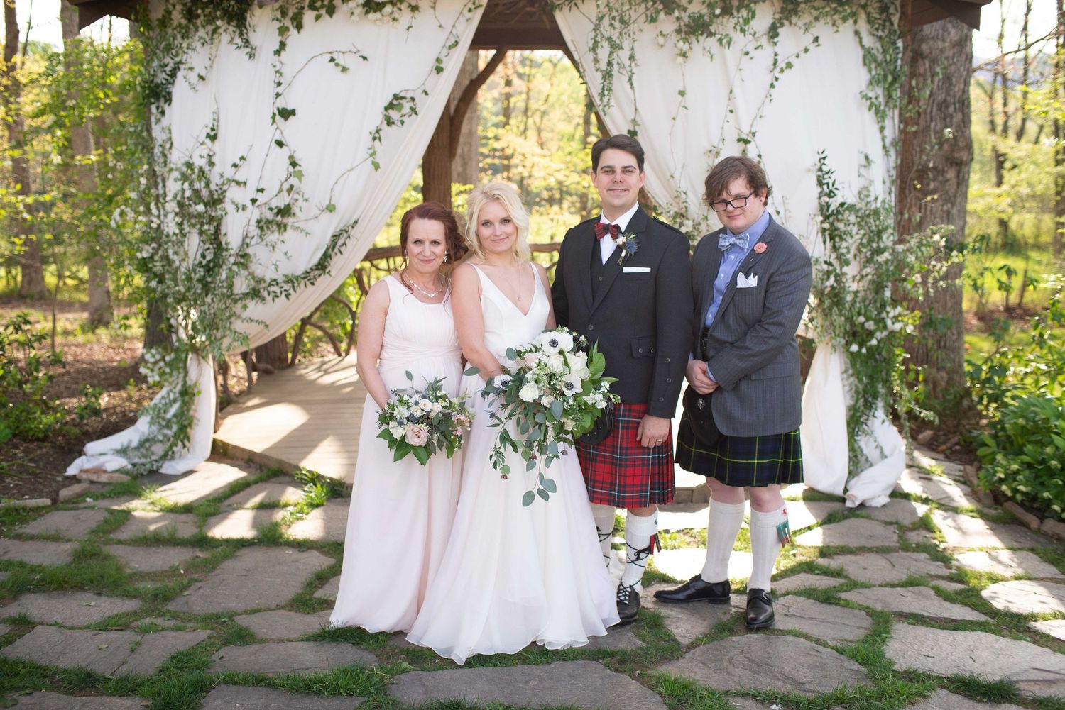 Wedding party poses under floral arch, bride in white gown, bridesmaid in pink, groom in suit, groomsman in tartan kilt.