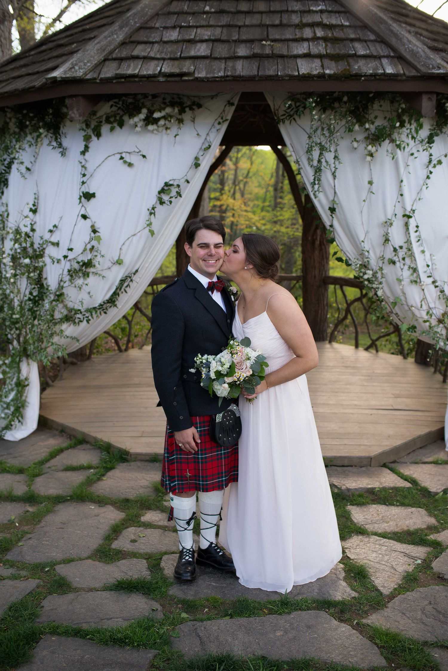 Bride and groom pose under decorated gazebo, groom wearing Scottish tartan kilt and bride in white strapless gown holding bouquet.