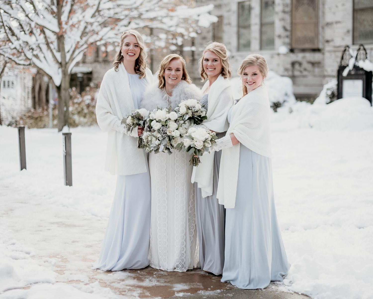 Bride and three bridesmaids in white wraps and light blue gowns pose outdoors in snowy winter wedding setting.