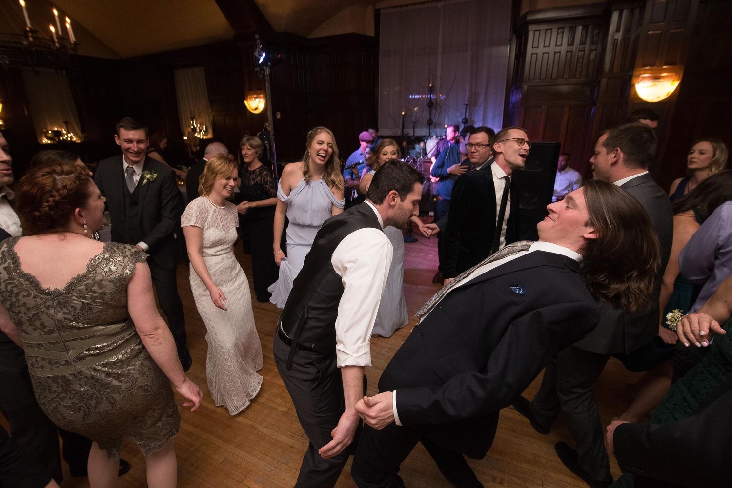 Guests dancing energetically at a wedding reception in an elegant wood-paneled ballroom with warm lighting.