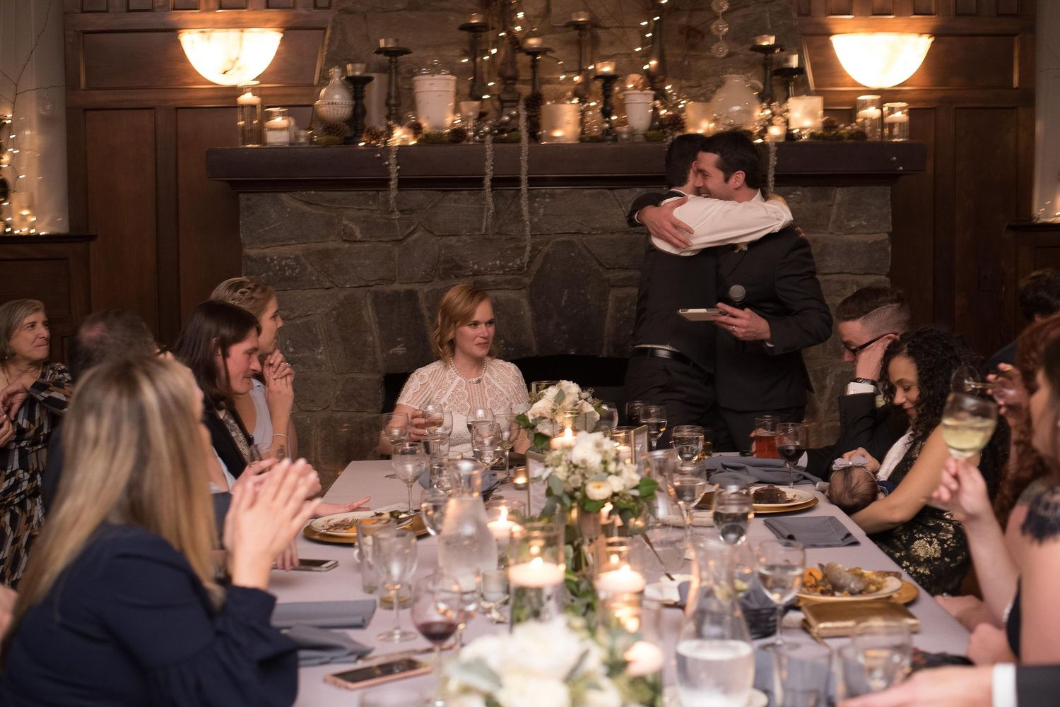 Guests celebrate at an elegant candlelit dinner party with floral centerpieces and a couple embracing near a fireplace.