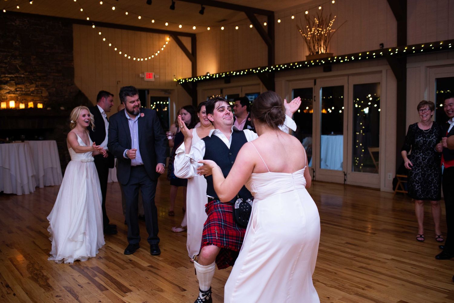 Guests dancing at a rustic wedding reception with string lights, a bride in white, and a man in a red kilt on the dance floor.