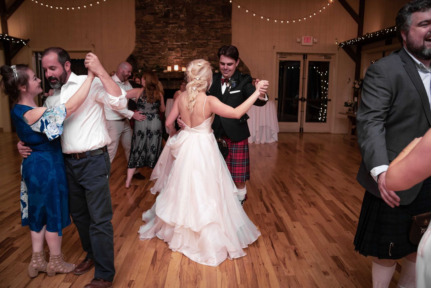 Bride and groom share first dance at rustic wedding reception venue with guests dancing around them.