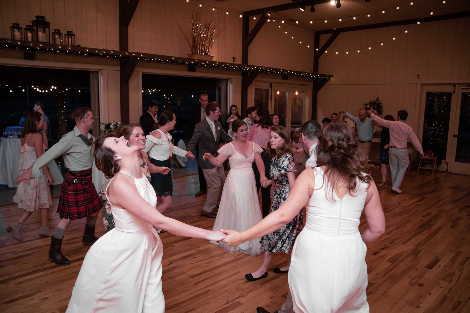 Brides in white dresses joyfully dance with wedding guests in a rustic barn venue adorned with string lights.