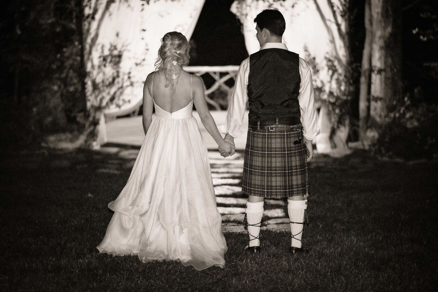 Black and white photo of bride and groom holding hands, groom wearing Scottish tartan kilt, walking toward rustic wooden arch.
