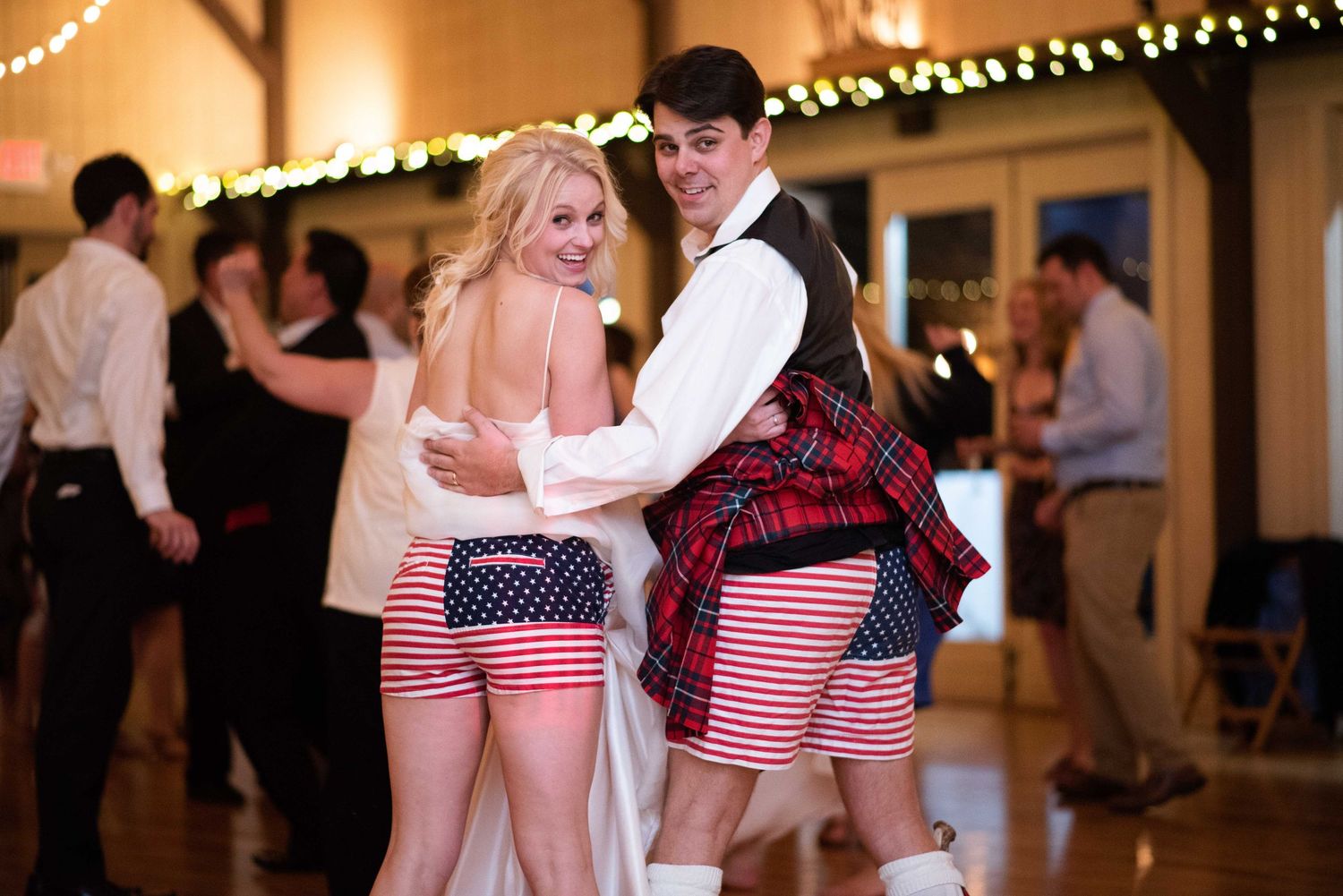 Couple dancing in patriotic American flag shorts and red tartan kilt at a festive barn wedding reception.