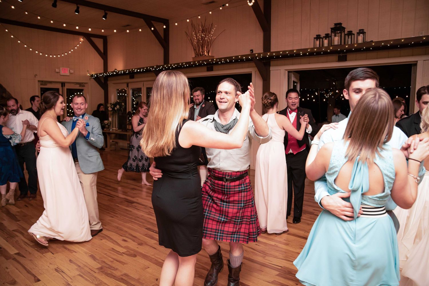 Guests dancing joyfully at a rustic barn wedding reception, with a woman in a plaid kilt standing out on the dance floor.