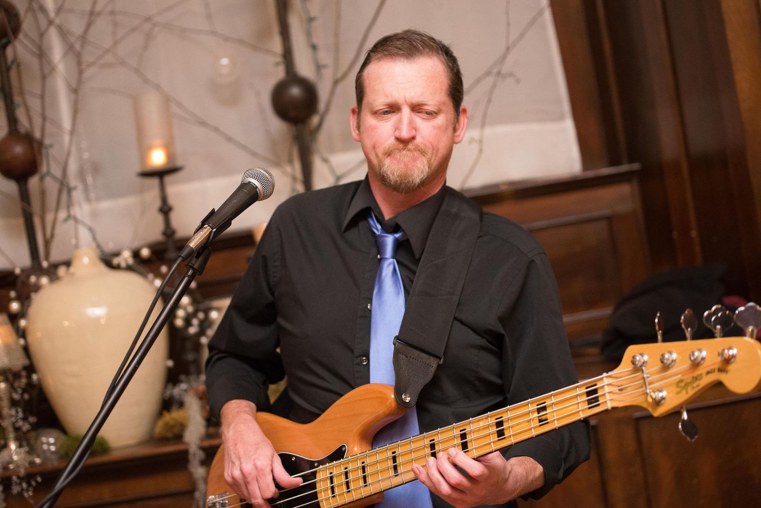 Man playing bass guitar at indoor event, wearing black shirt and blue tie, performing near microphone stand.