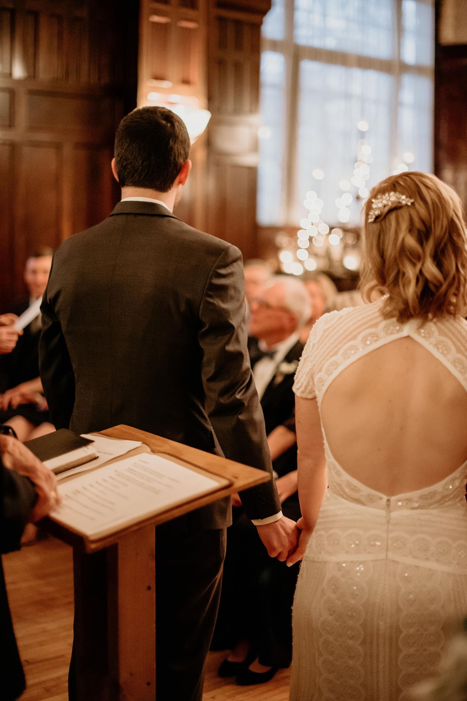 Bride and groom holding hands at wedding ceremony altar, bride in elegant backless lace gown with floral hair accessory.
