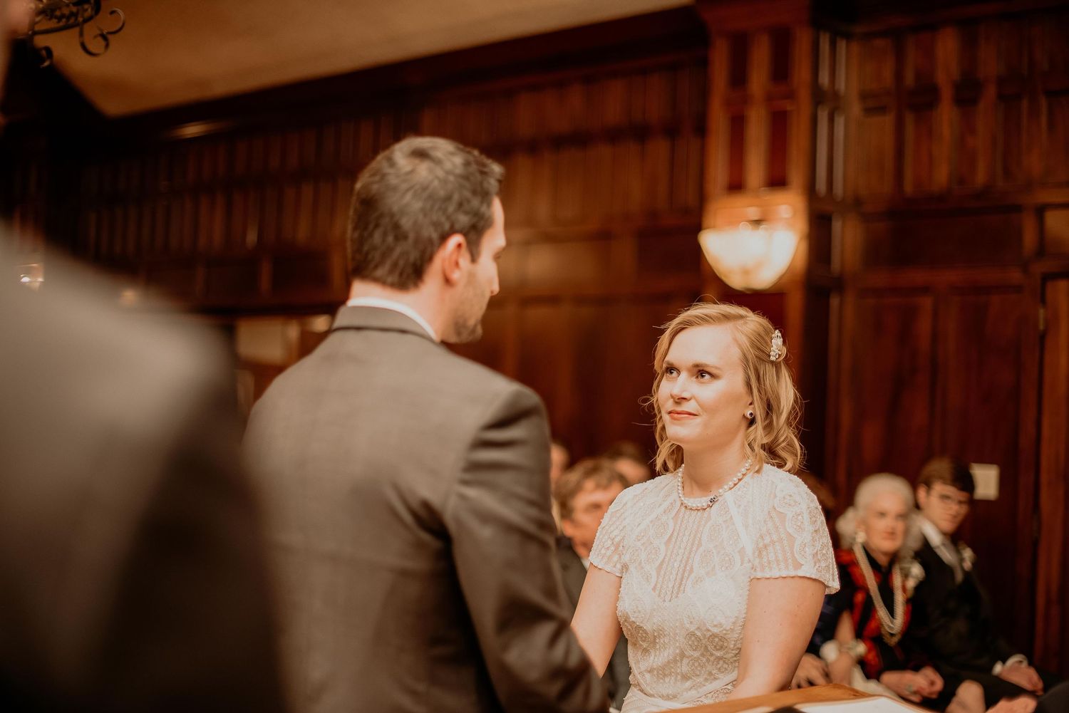Bride smiling at groom during intimate wedding ceremony in elegant wood-paneled venue with guests seated behind.